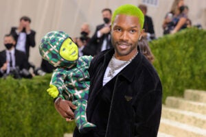 Frank Ocean smiling wearing a Prada jacket, skull and crossbones necklace and shaved green hair holding a green alien doll.